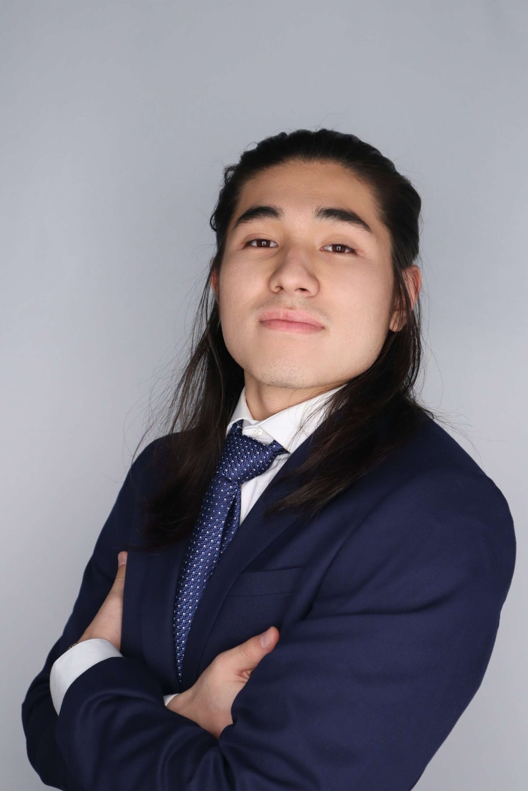 Student wearing a suit and tie poses with arms crossed on a gray background